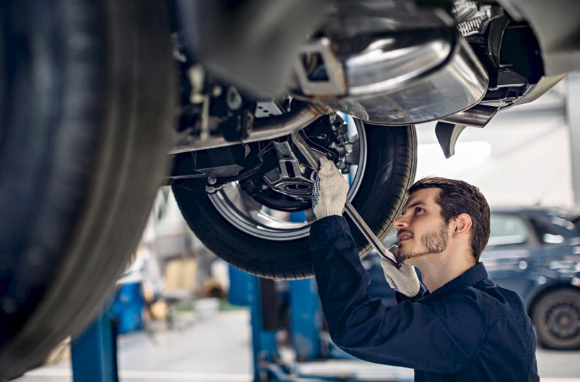 A mechanic in a garage is working under a vehicle, using a wrench to adjust or fix parts while looking up intently.