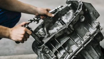 The image shows a person working on an automotive part, likely a gearbox or transmission, holding tools and performing maintenance or repairs.